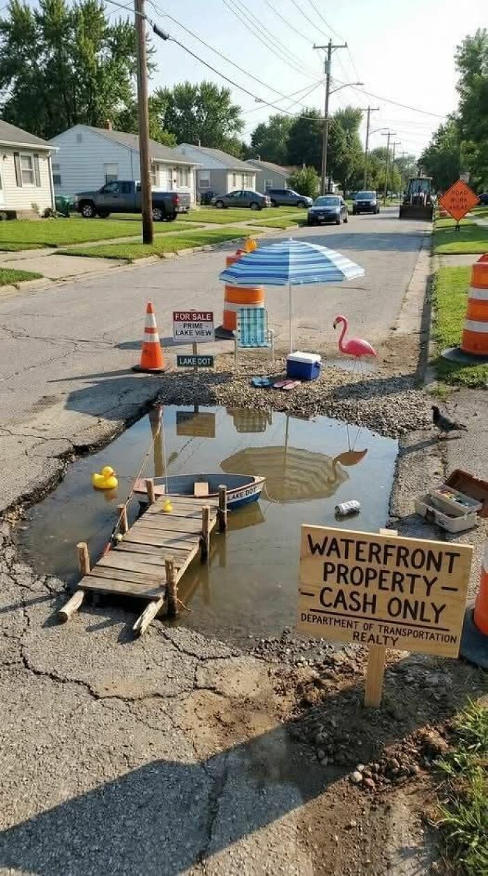 Humorous pothole turned into a miniature waterfront property with dock, boat, umbrella, and flamingo sign for laughs.