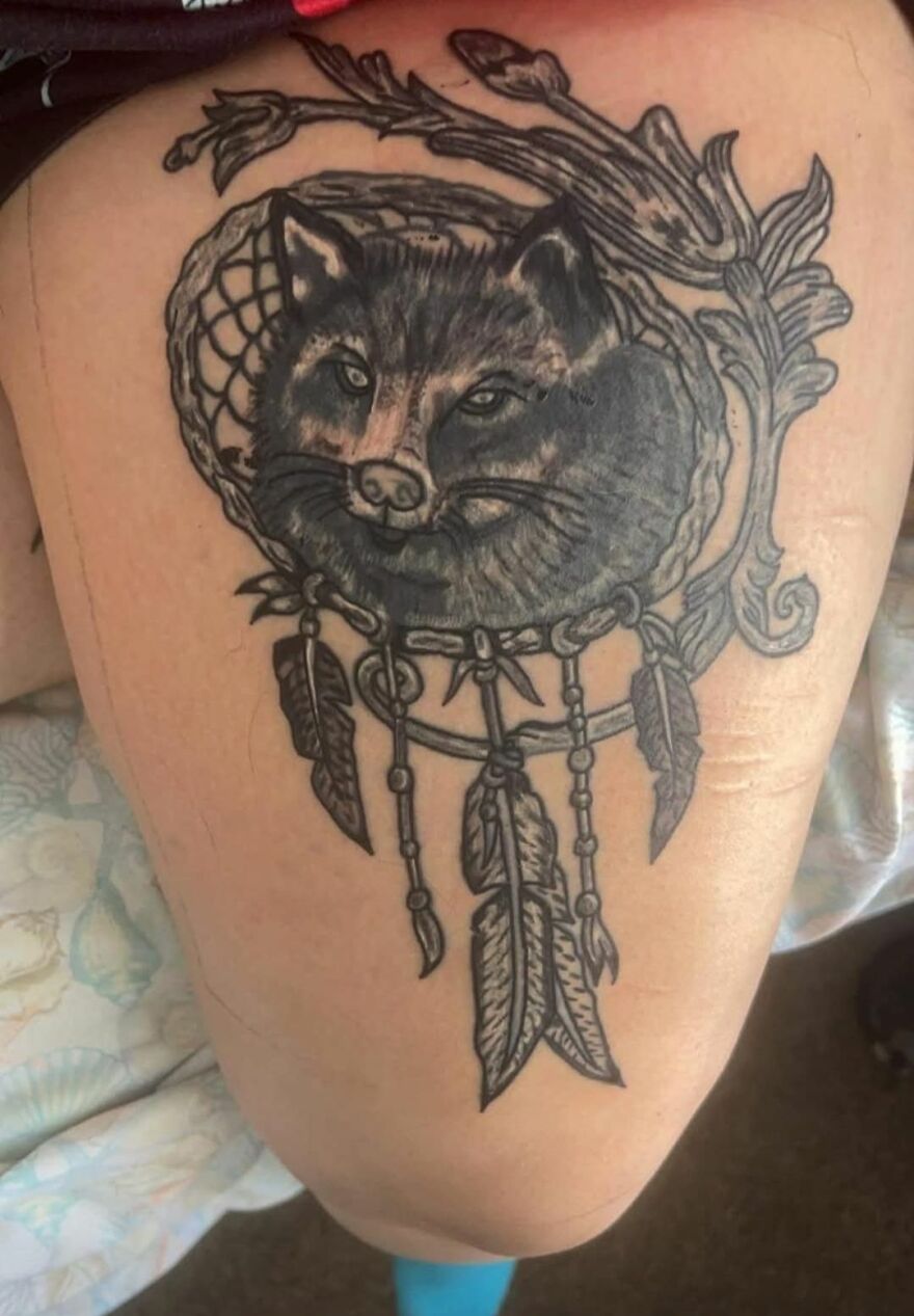 Black and gray tattoo of a fox inside a dreamcatcher with feathers on a person’s upper arm, showcasing unusual tattoo art.