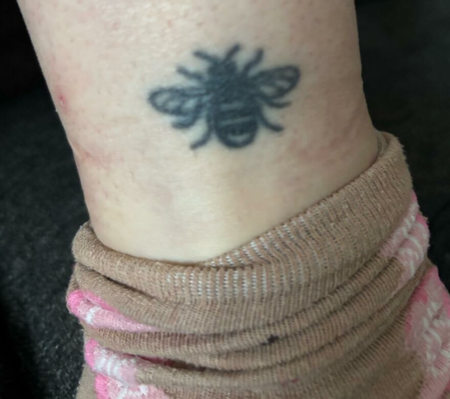 Small blurry bee tattoo on ankle above a pink and brown sock, an example of questionable tattoos and tattoo artists.