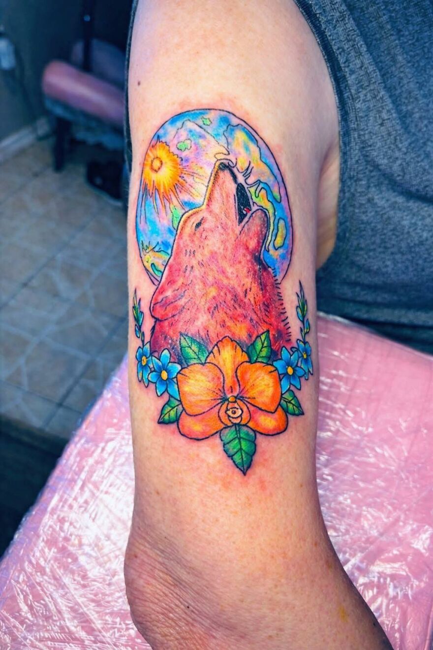 Colorful tattoo of a howling wolf with a sun and moon background, surrounded by orange and blue flowers on a forearm.