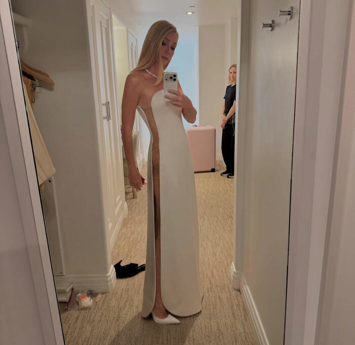 Woman in a sleek white risky Oscars dress with a high slit, posing for a mirror selfie in a hallway.