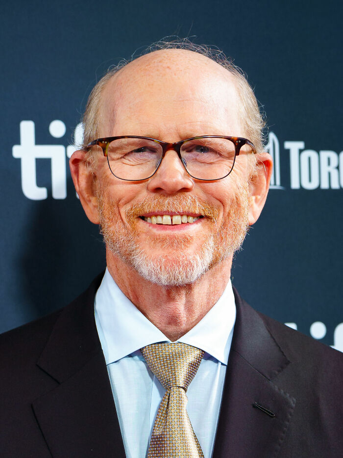 Ron Howard smiling at a film festival event, wearing glasses, suit, and patterned tie, highlighting his bio and career.