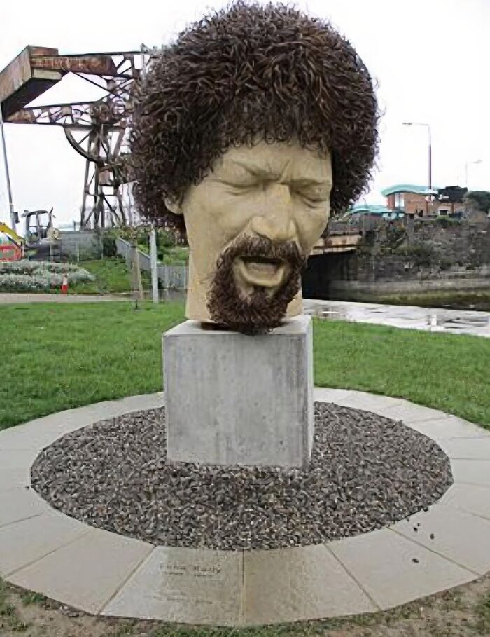 Large surreal statue of a man's head with curly hair and beard outdoors, part of statues from around the world that question reality