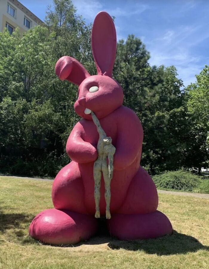 Large pink rabbit statue holding a small human figure in its mouth outdoors, one of the statues that make people question reality.