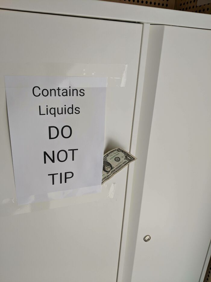 Sign reading contains liquids do not tip with a dollar bill placed sarcastically as a tip on a white cabinet.
