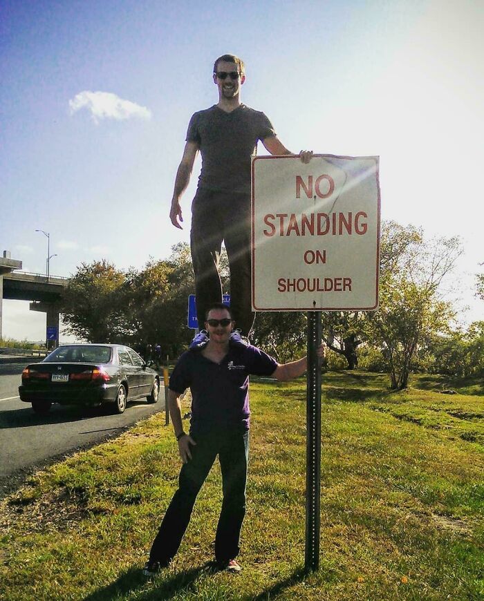 Two men humorously defying a no standing sign on a roadside shoulder, showcasing sarcastic and ironic behavior.