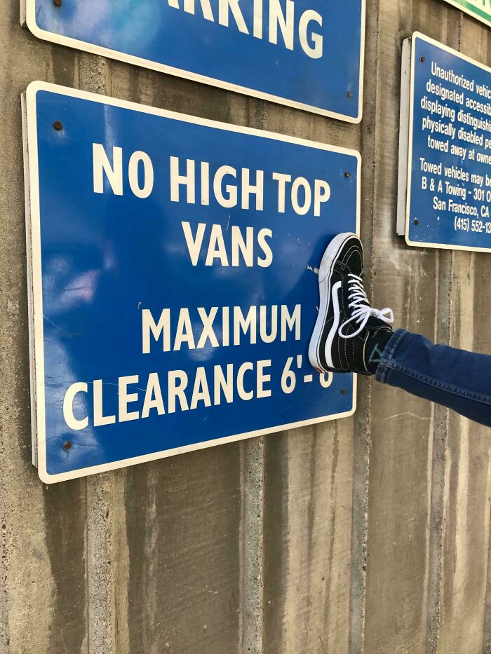 Blue parking sign reading no high top vans maximum clearance 6 feet with a person’s leg and sneaker playfully kicking it.