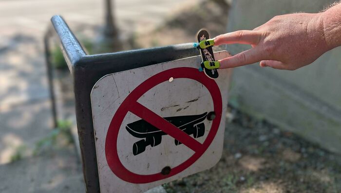 Hand holding a mini skateboard over a no skateboarding sign, showcasing a sarcastic and ironic moment.