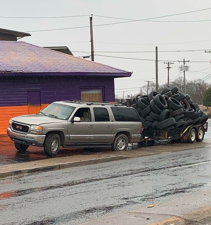Silver SUV towing a trailer overloaded with old tires parked by a purple and orange building on a rainy day, sarcastic image.
