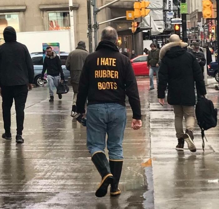 Man wearing a jacket with sarcastic text "I hate rubber boots" walking on a rainy city street, capturing ironic humor.