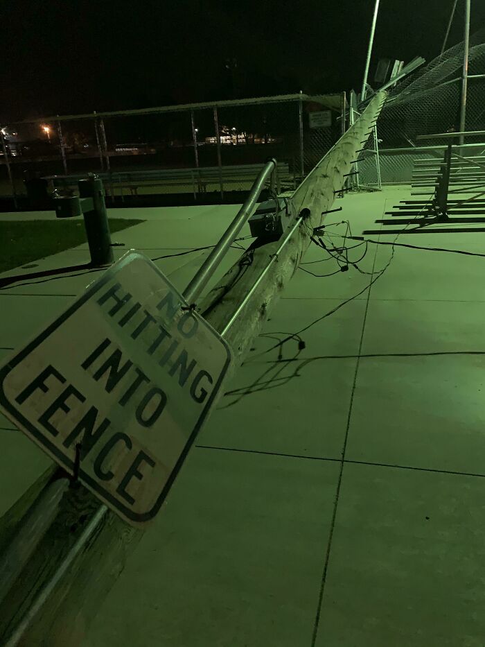 Fallen utility pole with sarcastic no hitting into fence sign lying on sidewalk at night, capturing ironic scene.