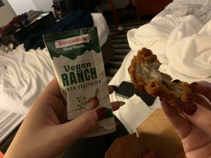Hand holding vegan ranch sauce packet and a bitten piece of fried chicken in a messy bedroom setting with sarcastic images vibe.