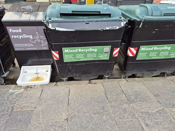 Outdoor recycling bins for food and mixed waste with a discarded sink placed beside them on a stone pavement.