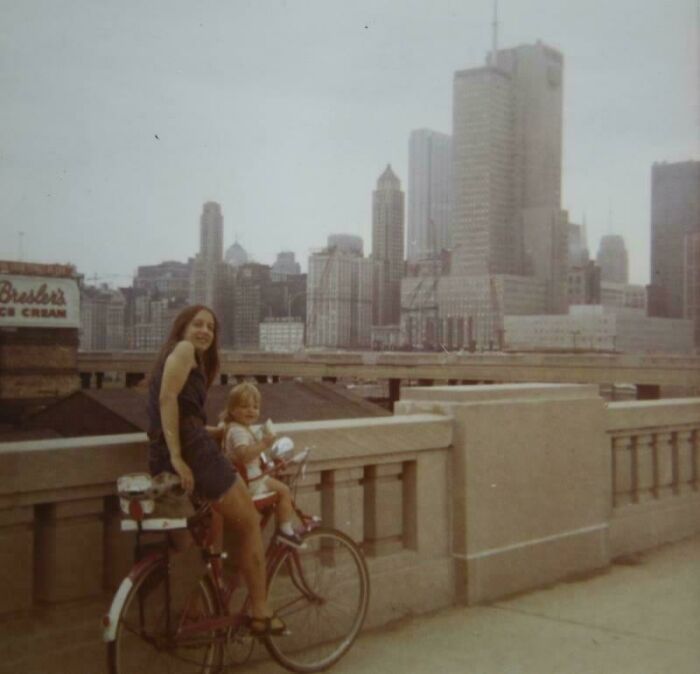 A candid old photo of a woman and child on a bike with a city skyline, revealing what the world looked like generations ago.