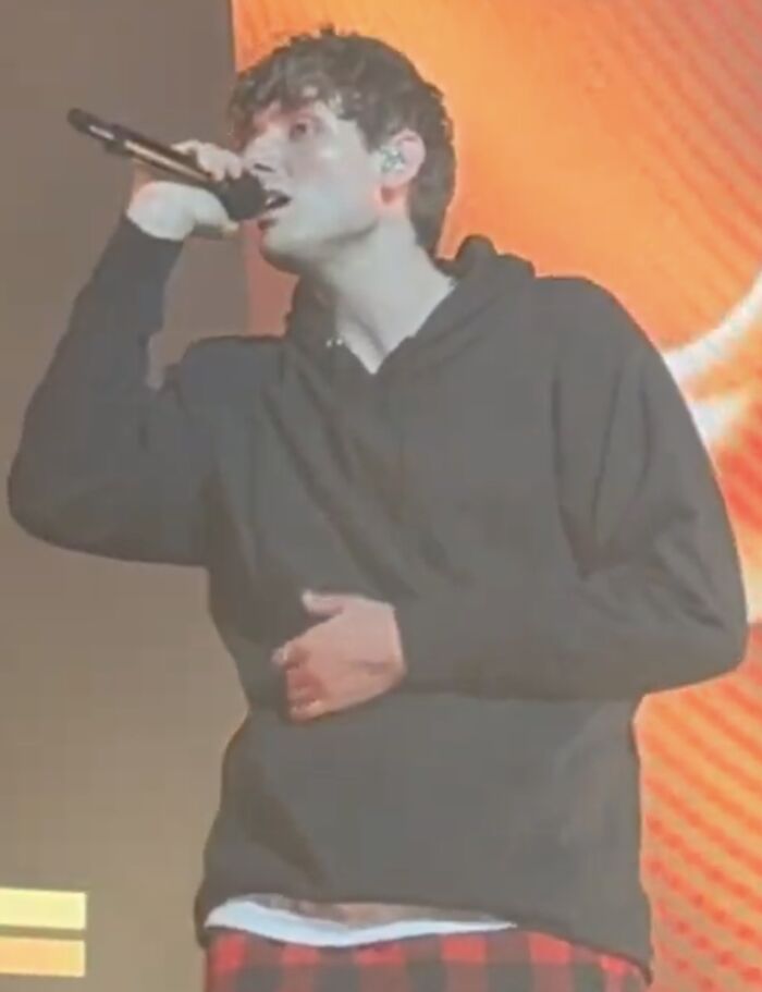 Jvke singing on stage wearing black hoodie and red plaid pants with bright orange background lighting.