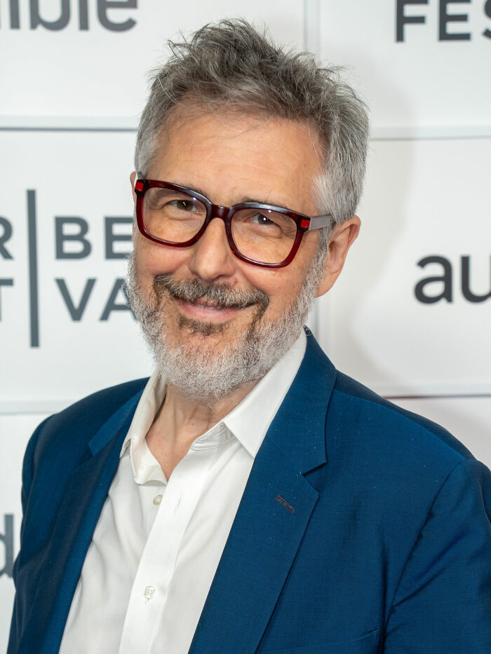 Ira Glass wearing glasses and a blue suit, smiling at an event, showcasing his bio and career highlights.