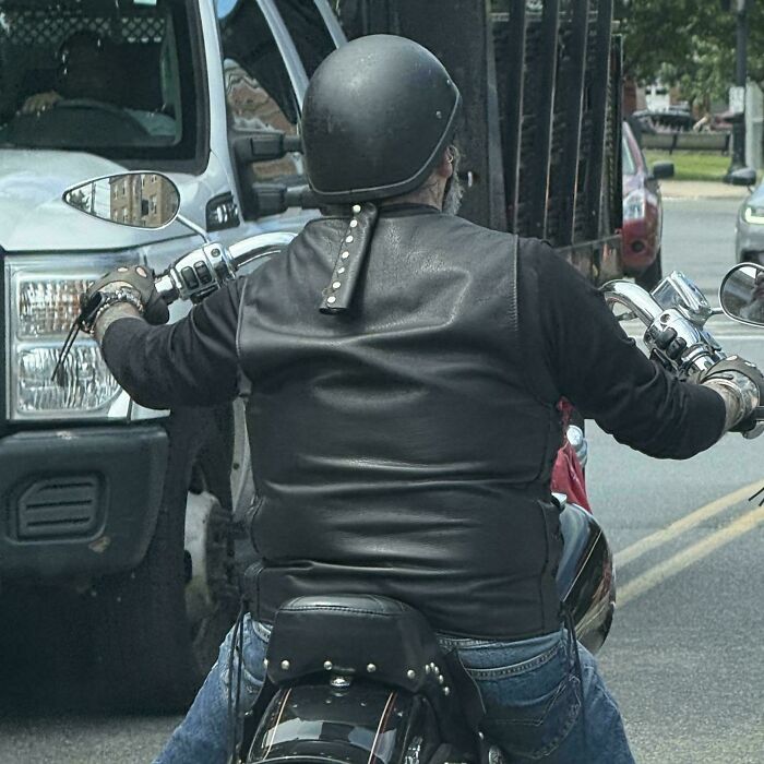 Biker wearing a black leather vest and helmet riding a motorcycle in traffic with unidentified objects nearby.