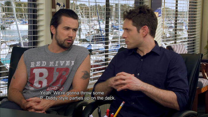 Two men sitting indoors with boats visible outside, captured in an old photo that aged badly for a facepalm moment.