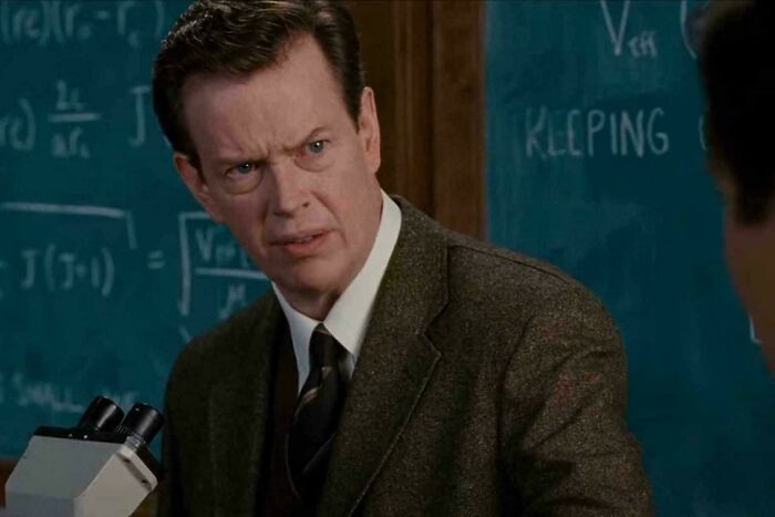 A man in a tweed jacket, white shirt, and dark tie with a confused expression, standing in front of a chalkboard, highlighting nonsensical movie details.