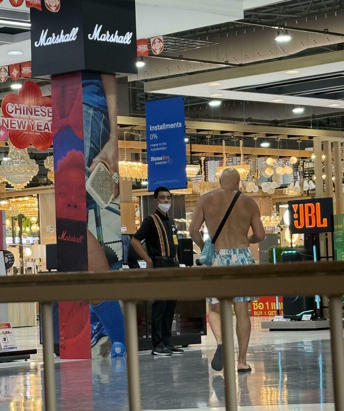 Shirtless tourist wearing shorts and sandals indoors, walking past a masked employee in a busy retail store, showcasing rude tourist behavior.