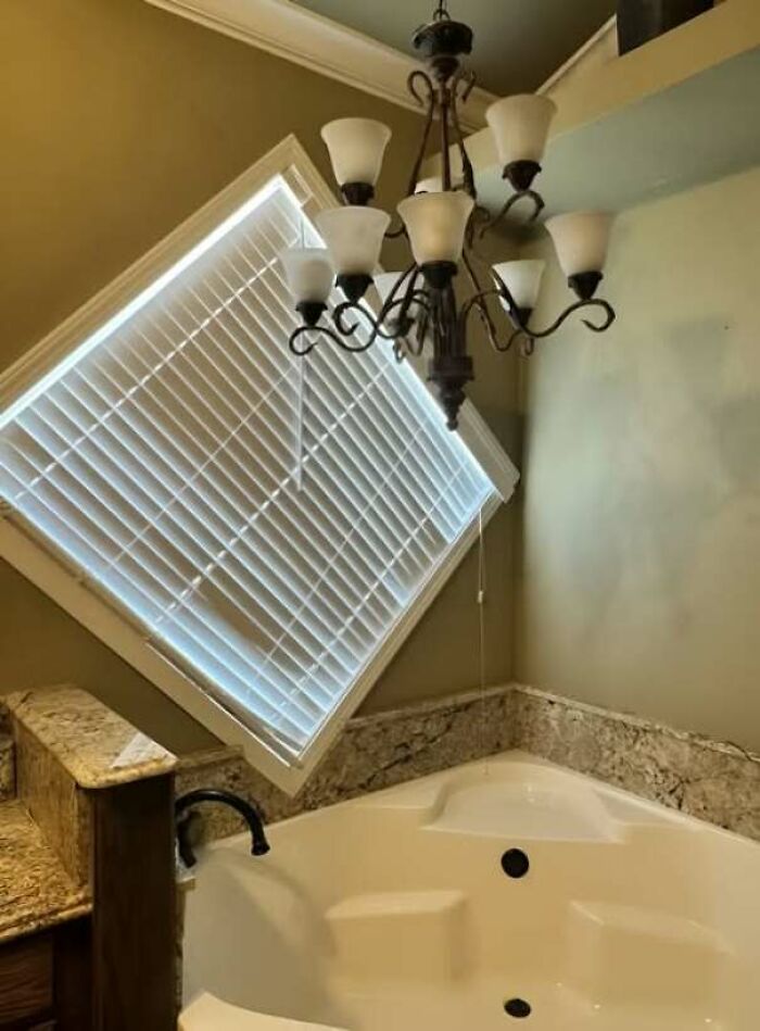 Bathtub under a crooked window with blinds and a chandelier in an abandoned or forgotten home interior.