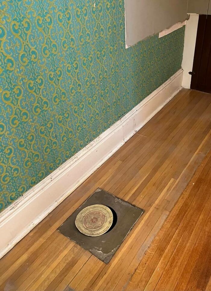 Floor vent cover replaced with a decorative plate in an abandoned home with worn wooden floors and vintage wallpaper.