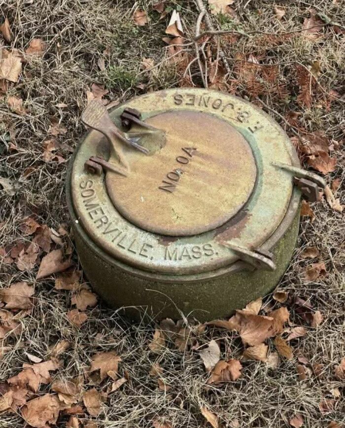 Rusty metal container labeled Somerville Mass, partially buried in grass and dead leaves, bizarre abandoned home find.