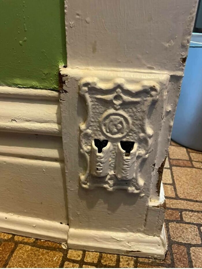 Old ornate white ceramic electrical outlet on peeling painted wall in an abandoned home with vintage flooring visible nearby
