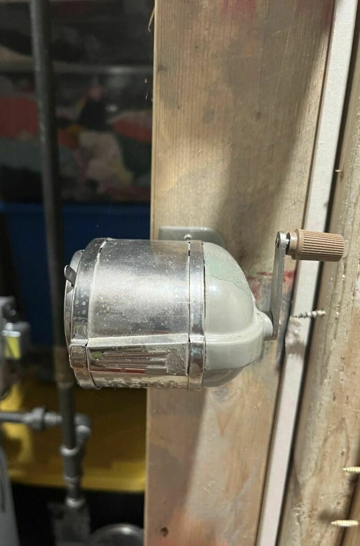 Old dusty pencil sharpener with hand crank mounted on a wooden wall in an abandoned or forgotten home.