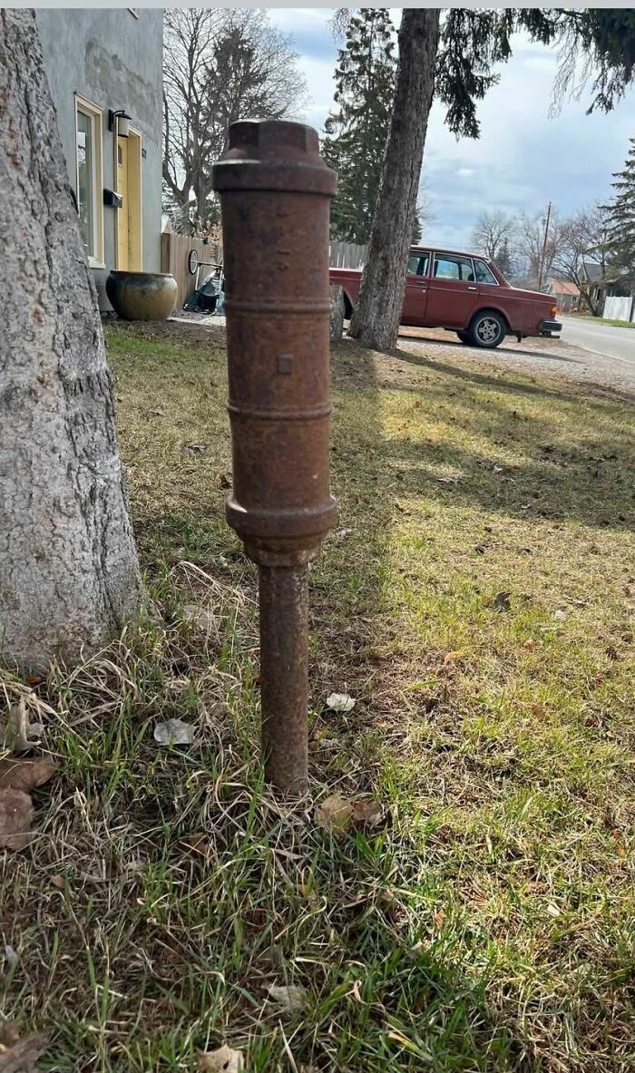 Rusty and weird metal pipe object partially buried in grass near an old house and vintage car in abandoned setting.
