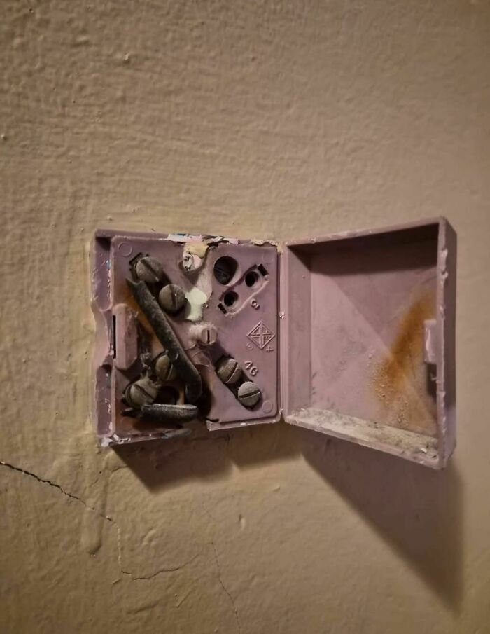 Old rusty electrical box with corroded screws inside a c*****d wall in an abandoned home setting.