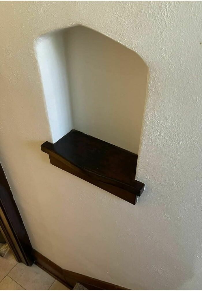 Small dark wooden shelf built into empty wall niche in an abandoned home with textured white walls and tiled floor.