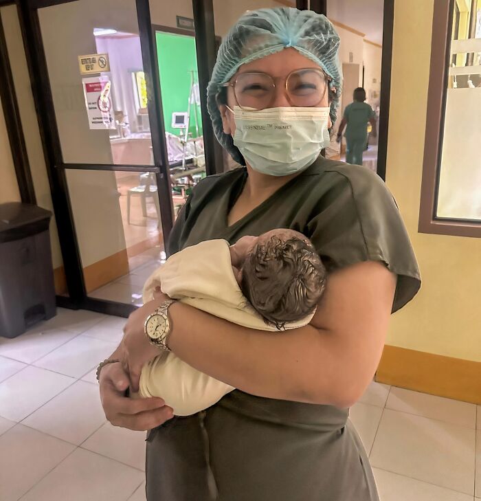 Female healthcare worker in scrubs and mask holding a newborn baby, representing ladies choosing lab coats over manicures.