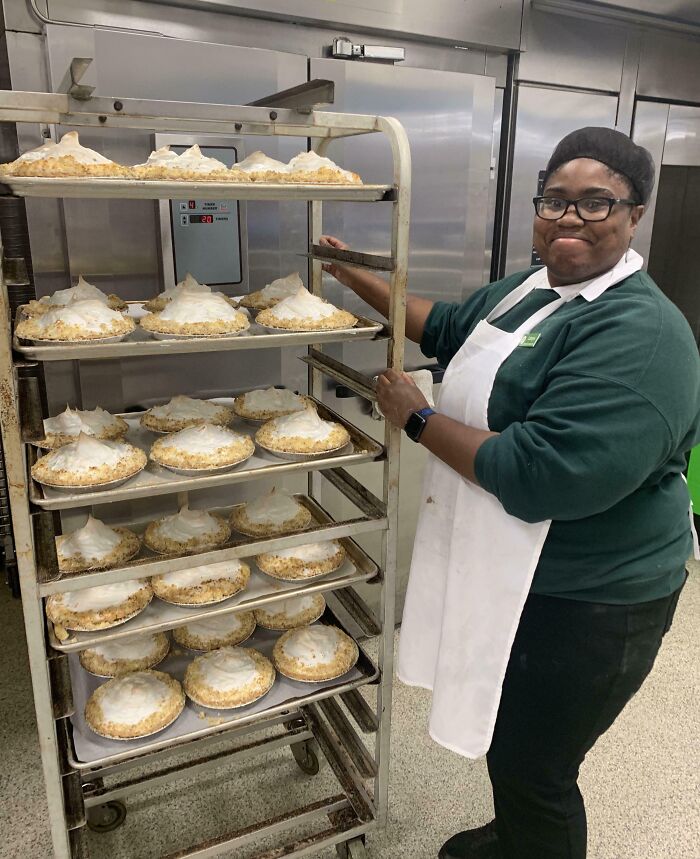 Woman in a lab coat and hairnet standing next to a rack filled with rows of freshly baked pies in a commercial kitchen.