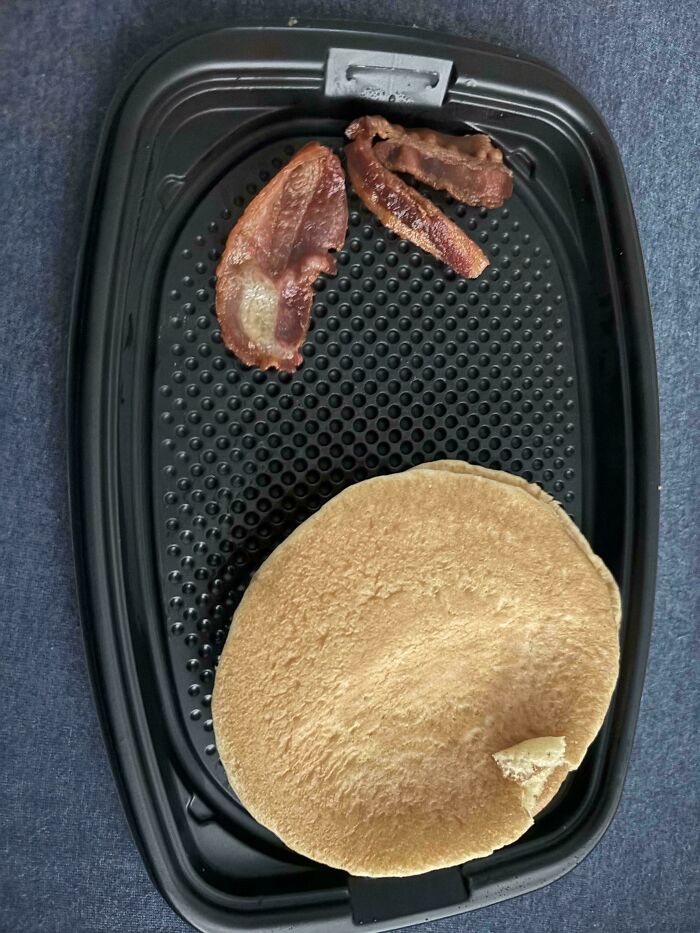 Small bacon strips and a large pancake served in a black plastic breakfast tray