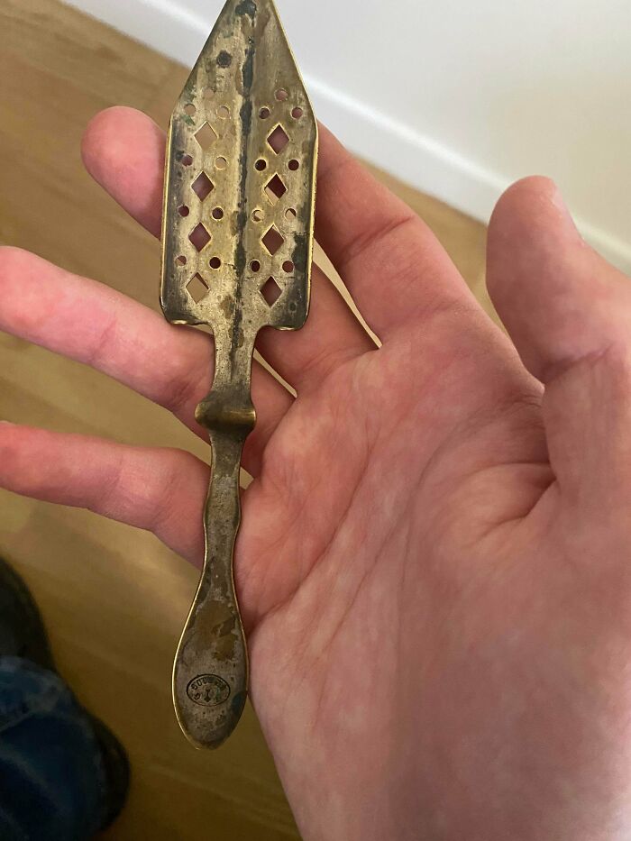 A hand holding a unique, remarkable antique, a brass absinthe spoon with diamond-shaped perforations.