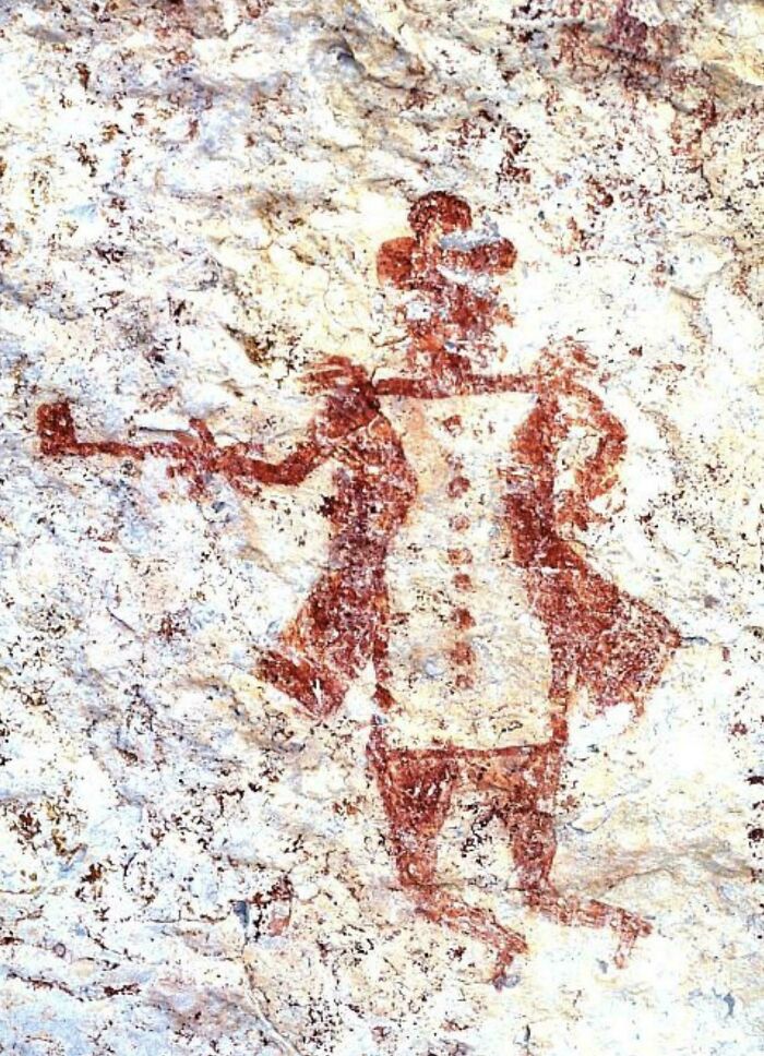 Prehistoric cave painting of humanoid figure with exaggerated features in red pigment