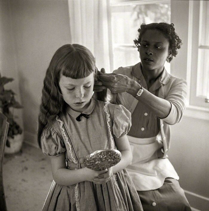 A woman combs a young girl's hair while the girl holds a mirror, an emotional historical photo of daily life.