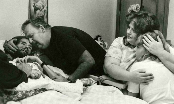 A man embraces a person in bed, surrounded by family, in this emotional historical photo capturing a poignant moment.