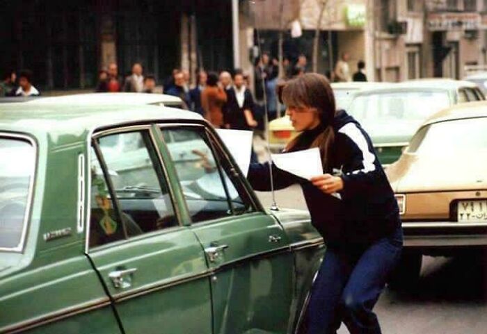 A woman in 70s attire leans into a green vintage car, holding papers in a street scene of historical photos.