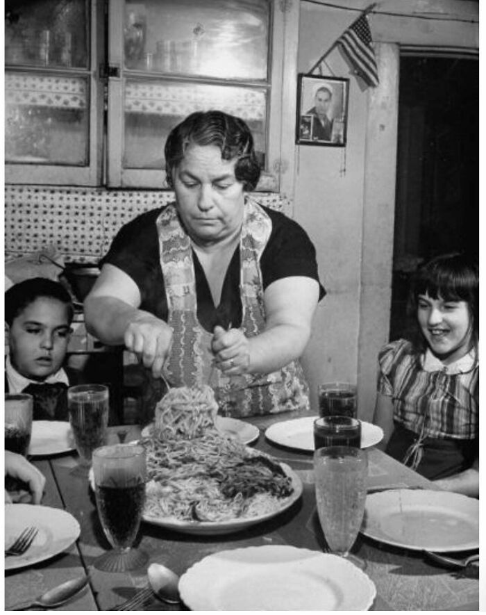 A fascinating historical photo of a woman serving a large dish of spaghetti to children at a dinner table.