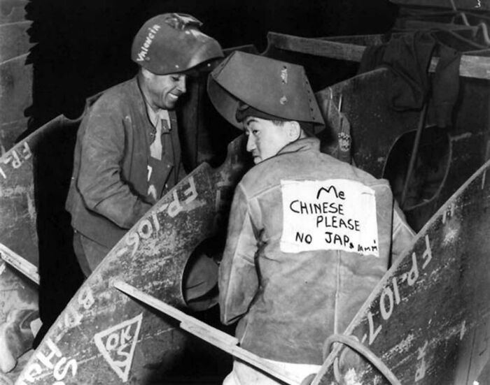 A historical photo of two welders, one with a sign saying Me Chinese Please, No J*p on his back, working.