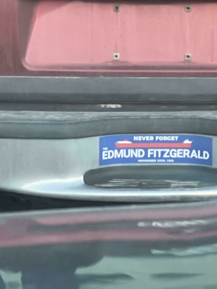 A close-up of a bumper sticker that reads Never Forget Edmund Fitzgerald on a red car, one of many funny bumper stickers.