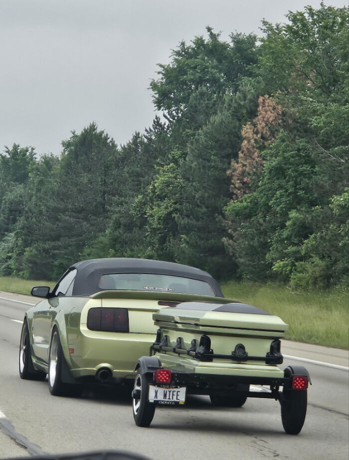 Green car towing matching green coffin trailer with funny license plate on highway