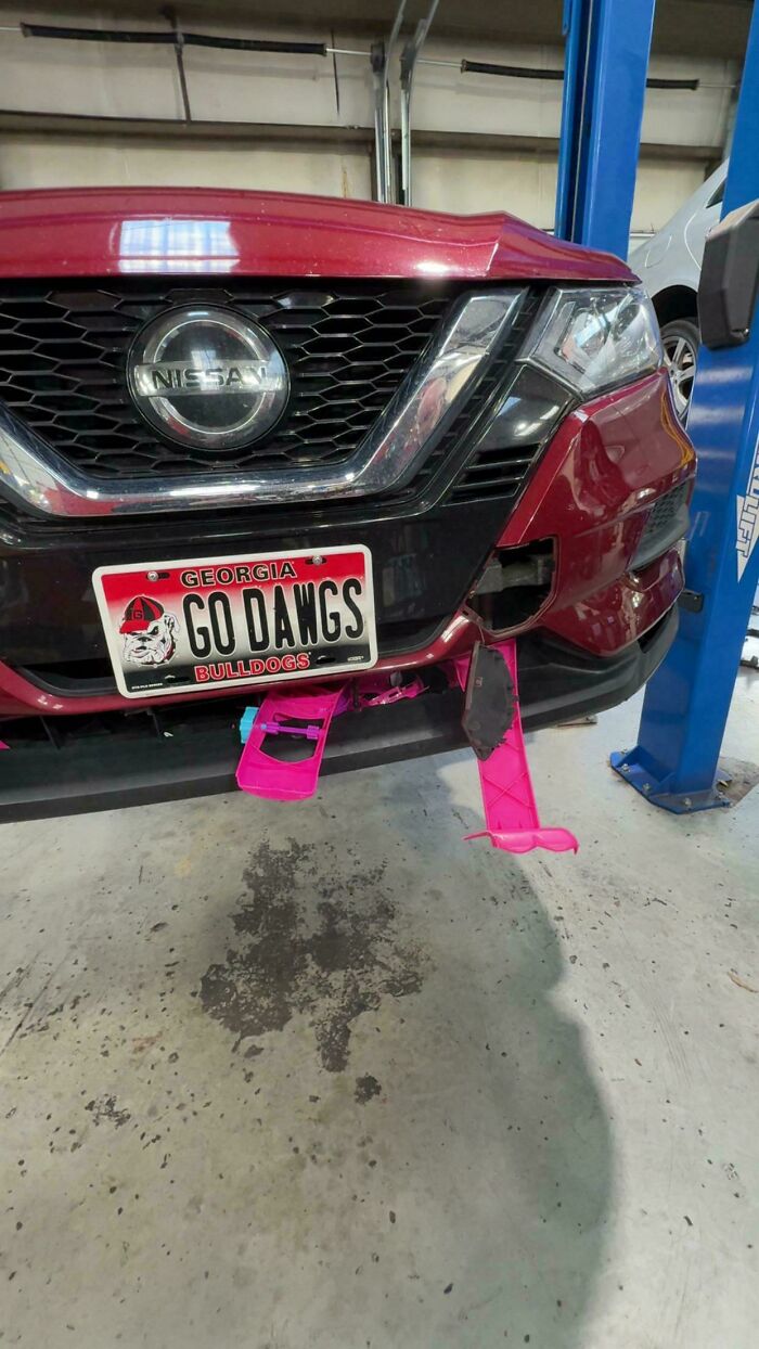 Front of a red Nissan car on a lift with a damaged bumper and pink streamers, a strange find for car mechanics.