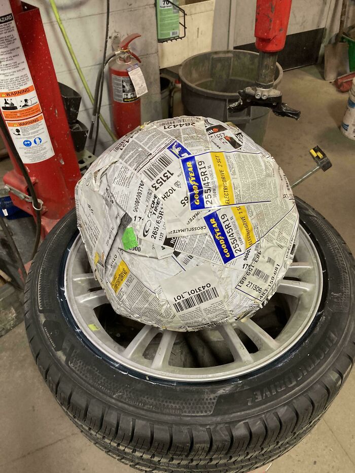 A car mechanic dealing with a car tire wrapped in newspaper, possibly for a prank or an unusual transport method.