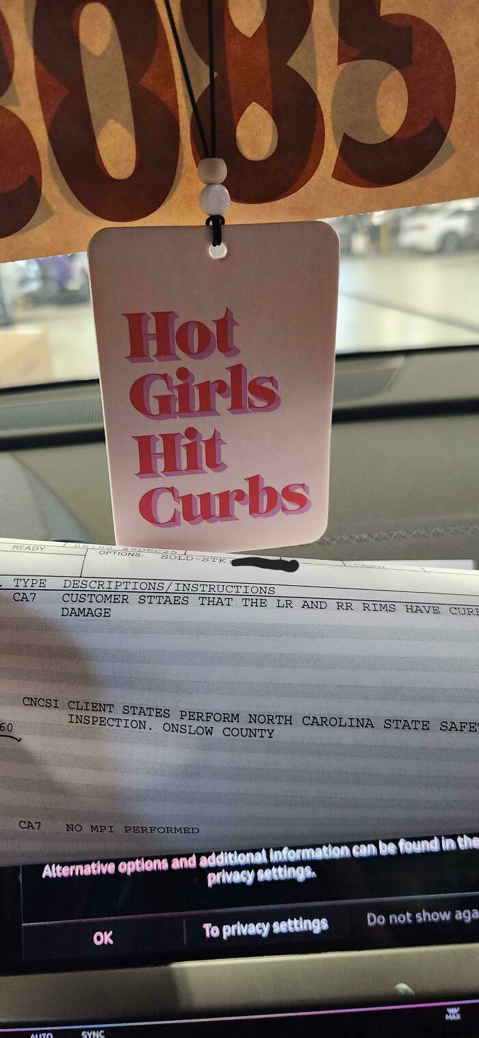 A car mechanic's humorous air freshener, Hot Girls Hit Curbs, hanging above car service notes.