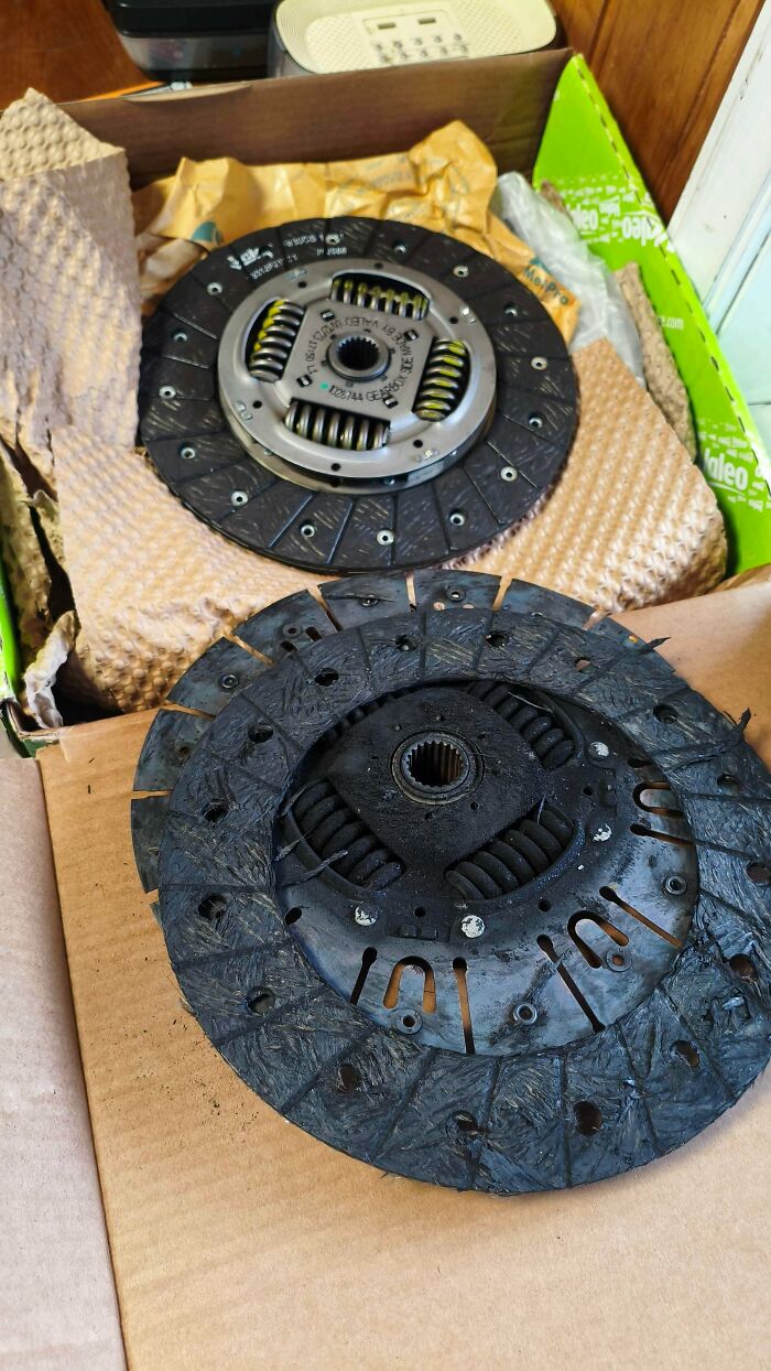 Car mechanics deal with a worn-out clutch plate next to a new one, showing the replacement needed.