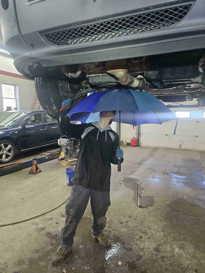 A car mechanic in dirty overalls holds an umbrella under a hoisted car, catching leaking fluid.