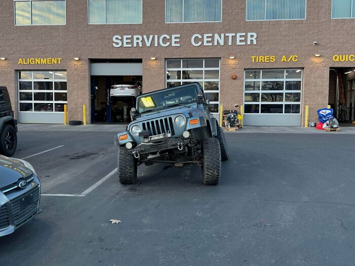 A gray Jeep with a severely damaged front wheel and suspension, parked askew outside a car service center.